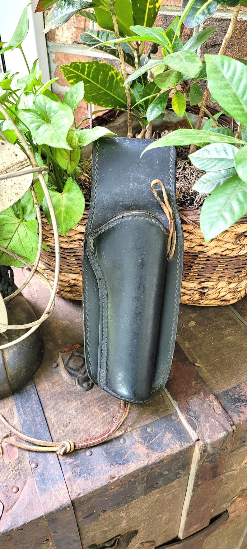 May include: Black leather holster with stitched detailing and a leather tie. The holster is placed on a vintage wooden trunk, with plants in the background. The holster is designed to hold a handgun.