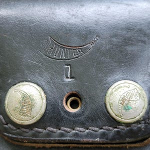 May include: Close-up of a black leather pouch with the word "HUNTER" embossed above the number "1". Two silver rivets with a horn design are visible. The leather has a stitched border.