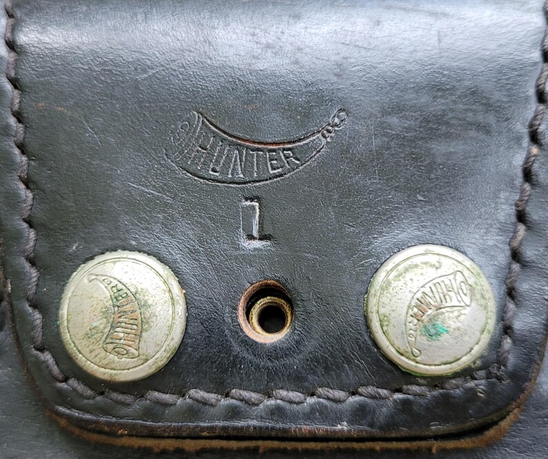 May include: Close-up of a black leather pouch with the word "HUNTER" embossed above the number "1". Two silver rivets with a horn design are visible. The leather has a stitched border.