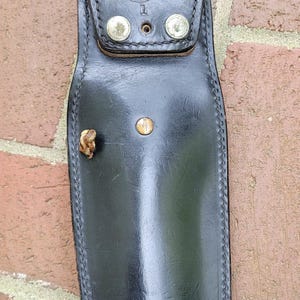 May include: Black leather sheath with a flap closure secured by two silver rivets. The word "HUNTER" is stamped on the flap. A small, decorative piece is attached to the side. The sheath is set against a brick wall.