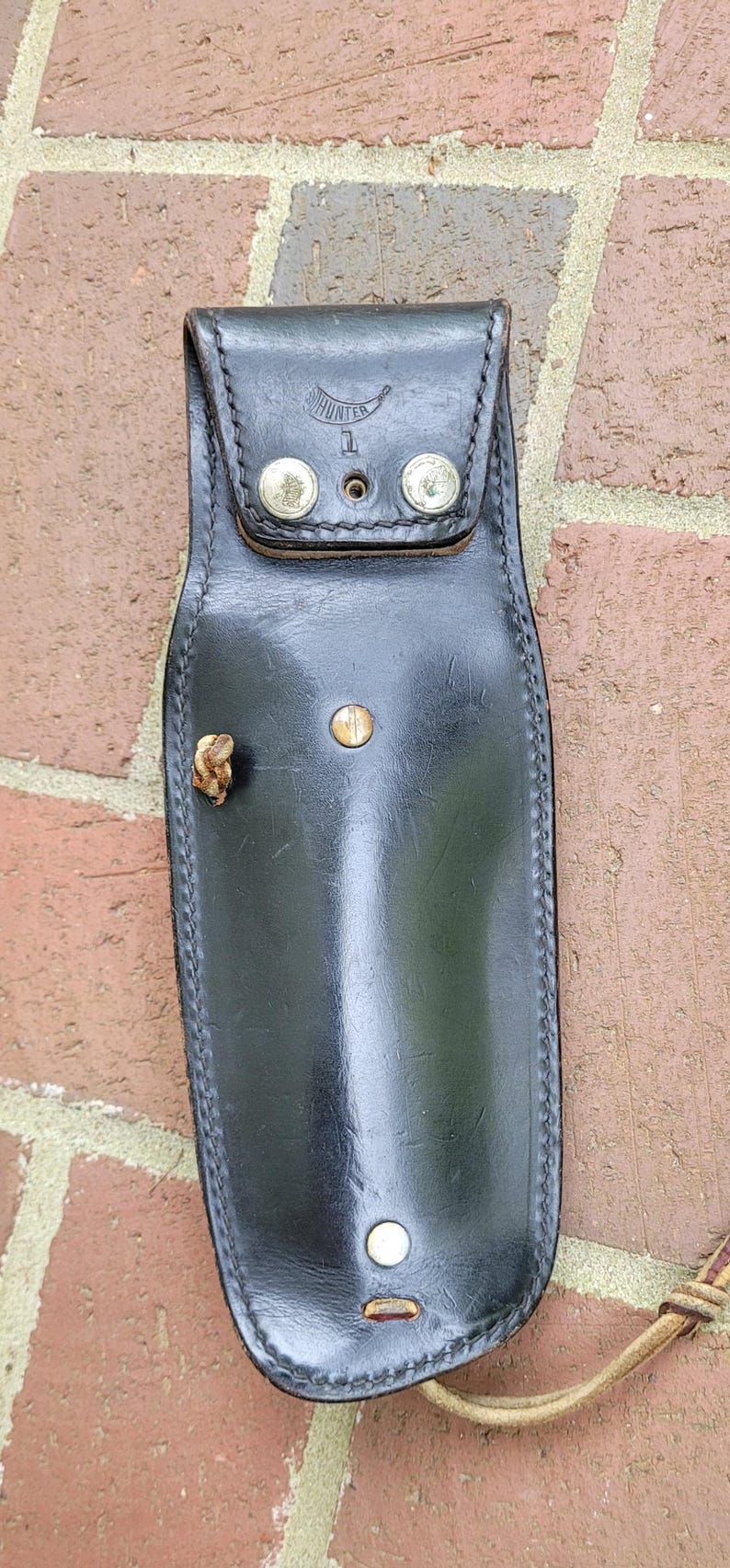 May include: Black leather sheath with a flap closure secured by two silver rivets. The word "HUNTER" is stamped on the flap. A small, decorative piece is attached to the side. The sheath is set against a brick wall.