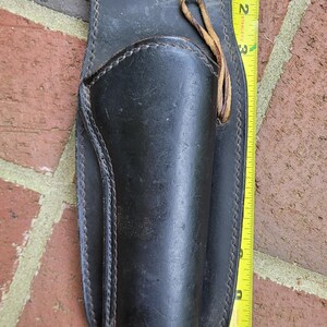 May include: Black leather holster with stitched edges and a leather tie. The holster is approximately 28 cm long. A yellow tape measure is placed next to the holster.