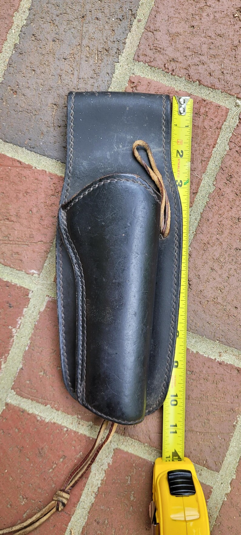 May include: Black leather holster with stitched edges and a leather tie. The holster is approximately 28 cm long. A yellow tape measure is placed next to the holster.