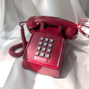 May include: A vintage burgundy push-button telephone with a handset and coiled cord. The phone has a rectangular base with a keypad of numbered buttons. The handset rests on top, and the cord is connected to the base.