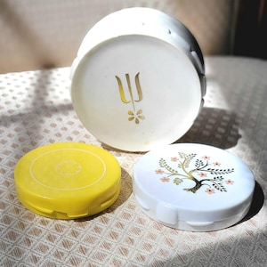 May include: Three vintage cosmetic compacts. One is white with a gold floral design, another is yellow, and the third is white with a gold emblem. The compacts are on a patterned surface.