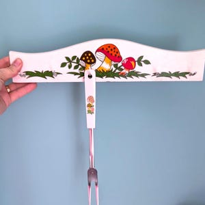 May include: A white, curved kitchen utensil holder with a pink handle and a metal fork. The holder is decorated with colorful mushroom illustrations and green foliage. The fork is attached to the holder by a pink handle with mushroom illustrations.