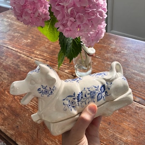 May include: A cream-colored ceramic butter dish shaped like a cow, featuring a delicate blue floral pattern across its body. The lid is sculpted in the form of a reclining cow with its head turned, and it rests on a rectangular base.
