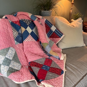 May include: A patchwork quilt featuring a variety of fabric squares in shades of pink, blue, red, and white. The quilt displays a repeating block pattern with intricate stitching throughout, finished with a solid cream-colored fabric border.