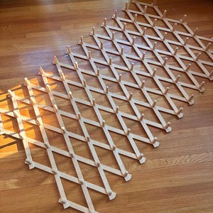 Könnte beinhalten: Ein ausziehbarer Wandgarderobenhalter aus hellem Holz mit Gitterdesign. Die Struktur besteht aus sich kreuzenden Holzleisten, die ein Rautenmuster bilden, wobei an jedem Verbindungspunkt kleine zylindrische Stifte angebracht sind.