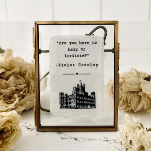 May include: A small, rectangular brass picture frame holds a white paper with the quote "Are you here to help or irritate?" by Violet Crawley. Below the quote is a black illustration of a castle. The frame is on a white surface.