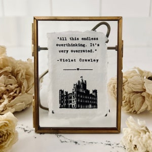 May include: A framed print featuring a quote by Violet Crawley from the TV show Downton Abbey. The quote reads "All this endless overthinking. It's very overrated." The print also includes a black and white illustration of Downton Abbey.