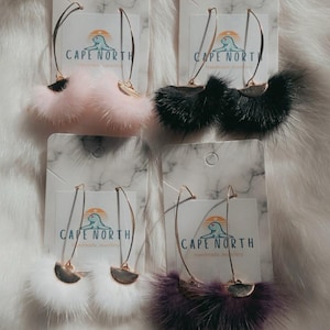 Può includere: Quattro paia di orecchini su cartoncini espositivi con motivo a marmo etichettati Cape North. Ogni paio presenta un arco in metallo dorato e un soffice inserto in pelliccia semicircolare in rosa chiaro, nero, bianco o viola scuro. Il testo recita Follow on FB for a chance to win a pair.