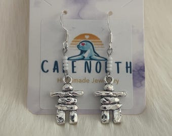 May include: A pair of silver-toned inukshuk dangle earrings featuring small white beads on the ear wires. The earrings are displayed on a white card with the text CAPE NORTH Handmade Jewelry and a logo of a seal under a sunset.