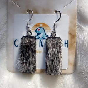 May include: A pair of rectangular drop earrings featuring silver-toned metal frames filled with textured, grey-toned animal fur. The earrings hang from silver-toned metal hooks and are displayed on a white card with a logo of a seal and the text CANH.