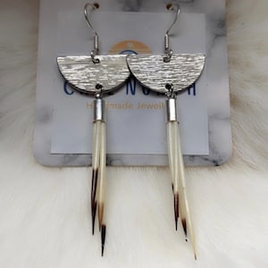 May include: Dangle earrings featuring a silver-toned semi-circular top piece with a textured finish, connected to a cluster of three pointed, off-white and brown quills. The earrings hang from silver-toned hooks and are displayed on a branded card.