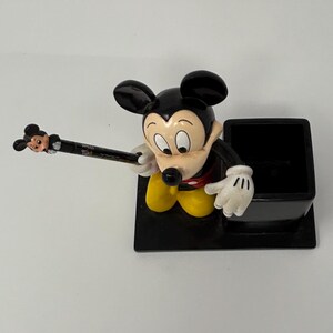 May include: A black pen holder featuring a Mickey Mouse figurine. The pen holder has a black base with a black square pen slot. Mickey Mouse is wearing yellow pants, white gloves, and a red shirt. A pen with a Mickey Mouse head is in his hand.