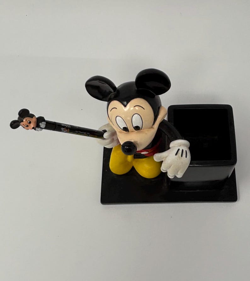May include: A black pen holder featuring a Mickey Mouse figurine. The pen holder has a black base with a black square pen slot. Mickey Mouse is wearing yellow pants, white gloves, and a red shirt. A pen with a Mickey Mouse head is in his hand.