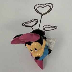 May include: A Minnie Mouse figurine with a black head, pink bow, and blue dress. The figurine holds three silver heart-shaped wire holders, perfect for displaying photos or notes. The figurine is approximately 4 inches tall.