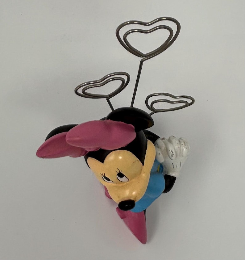 May include: A Minnie Mouse figurine with a black head, pink bow, and blue dress. The figurine holds three silver heart-shaped wire holders, perfect for displaying photos or notes. The figurine is approximately 4 inches tall.
