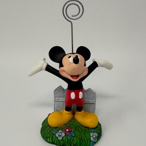 May include: A Mickey Mouse figurine with arms outstretched, wearing red shorts, yellow shoes, and white gloves. The figure stands on a green base with flowers and a small picket fence. A silver wire holds a photo or note.