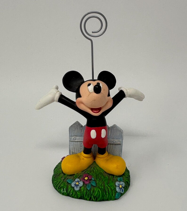 May include: A Mickey Mouse figurine with arms outstretched, wearing red shorts, yellow shoes, and white gloves. The figure stands on a green base with flowers and a small picket fence. A silver wire holds a photo or note.