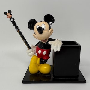 May include: A Mickey Mouse desk organizer featuring a black square pen holder and a figurine of Mickey Mouse. Mickey is wearing red shorts, black shoes, and white gloves. A black pencil with a Mickey Mouse topper is in his hand.