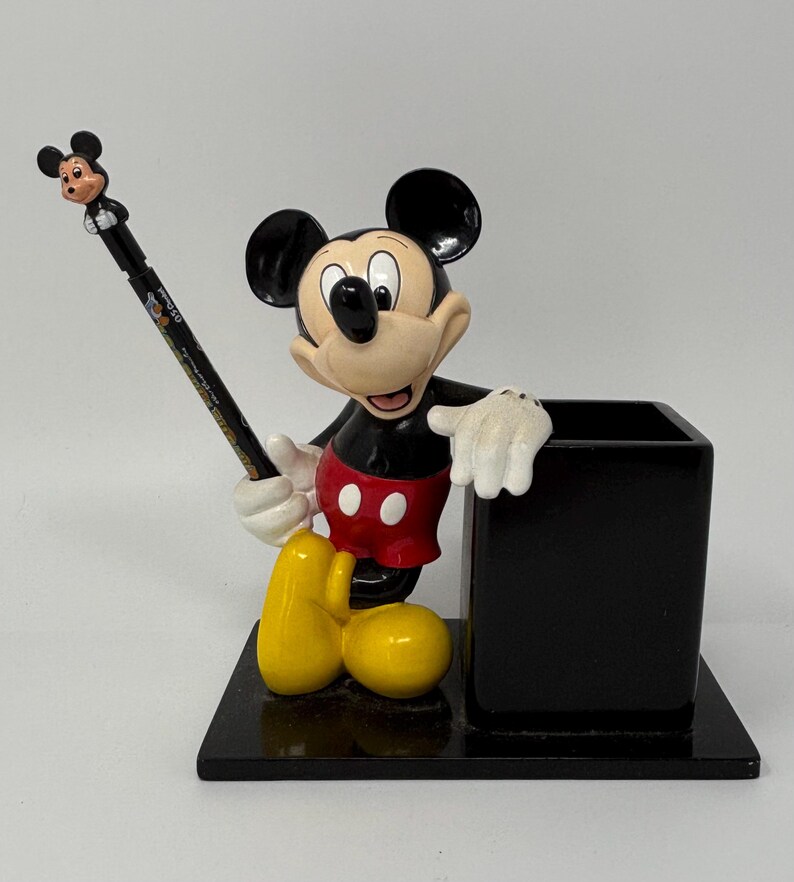 May include: A Mickey Mouse desk organizer featuring a black square pen holder and a figurine of Mickey Mouse. Mickey is wearing red shorts, black shoes, and white gloves. A black pencil with a Mickey Mouse topper is in his hand.