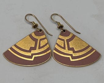 May include: A pair of gold-toned earrings with a fan-like shape. The earrings feature a mauve background with a gold geometric design. The ear wires are gold-colored. The earrings are displayed on a white surface.