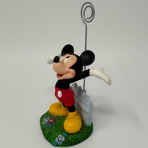 May include: A Mickey Mouse figurine with a silver wire holder. Mickey is in his classic outfit: red shorts, yellow shoes, black body, and white gloves. He stands on a green base with flowers and a white picket fence.