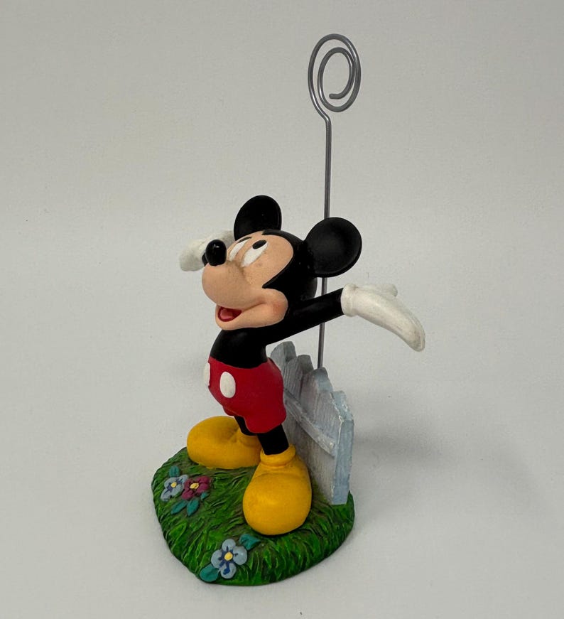 May include: A Mickey Mouse figurine with a silver wire holder. Mickey is in his classic outfit: red shorts, yellow shoes, black body, and white gloves. He stands on a green base with flowers and a white picket fence.
