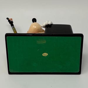 May include: A Goofy figurine, featuring a tan head, black ears, and white gloves, is seated behind a black desk. A pencil with a colorful design rests on the desk. The base is green with a black border.