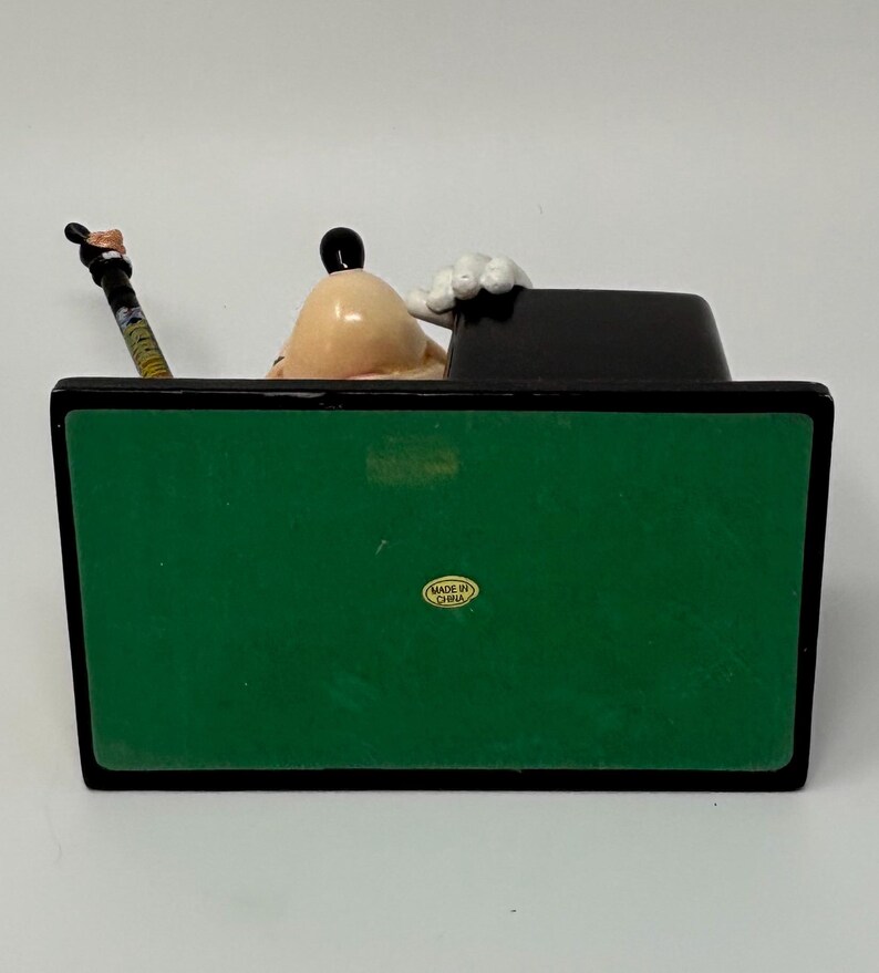 May include: A Goofy figurine, featuring a tan head, black ears, and white gloves, is seated behind a black desk. A pencil with a colorful design rests on the desk. The base is green with a black border.