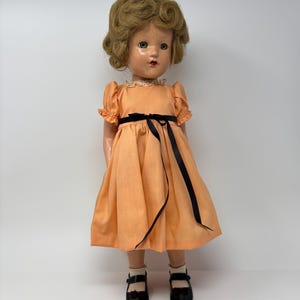 May include: A vintage doll with blonde curly hair, wearing an orange dress with a black ribbon and black shoes. The doll has a lace collar and is standing against a white background. The doll's features include painted eyes and red lips.