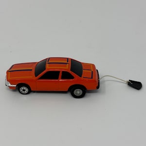 May include: Orange toy car with black windows and a pull string. The car has orange, purple, and yellow stripes on the roof and hood. The wheels are black with silver rims. The pull string has a black handle.