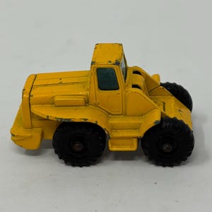 May include: A yellow toy construction vehicle with black wheels. The vehicle has a cab with a windshield and a large scoop in the front.