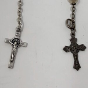 May include: Close-up of two rosaries. One rosary features a silver crucifix with a detailed depiction of Jesus Christ. The other rosary has a dark metal crucifix with a similar depiction. Both rosaries have a chain and are set against a white background.