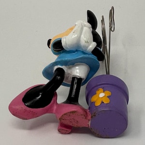 May include: A Minnie Mouse figurine in a blue dress and pink shoes, holding a purple flower pot with a yellow flower. The figurine has black ears and a white glove. The pot has a metal clip.