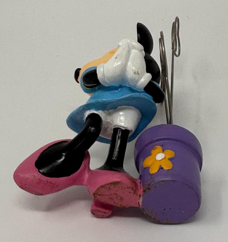 May include: A Minnie Mouse figurine in a blue dress and pink shoes, holding a purple flower pot with a yellow flower. The figurine has black ears and a white glove. The pot has a metal clip.