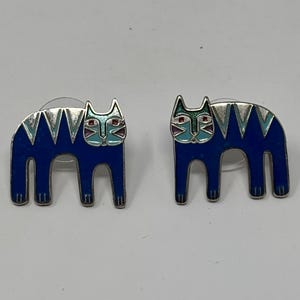 May include: A pair of cat-shaped stud earrings. Each earring features a blue enamel cat design with silver-colored outlines and details. The cat's face has a teal and red color scheme.