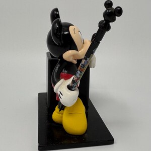 May include: A black Mickey Mouse figurine holding a pen with a colorful design. The figurine has yellow shoes, white gloves, and a red shirt. The pen has a black top with three circles. The figurine is on a black rectangular base.