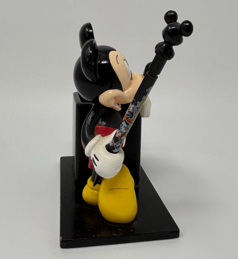 May include: A black Mickey Mouse figurine holding a pen with a colorful design. The figurine has yellow shoes, white gloves, and a red shirt. The pen has a black top with three circles. The figurine is on a black rectangular base.