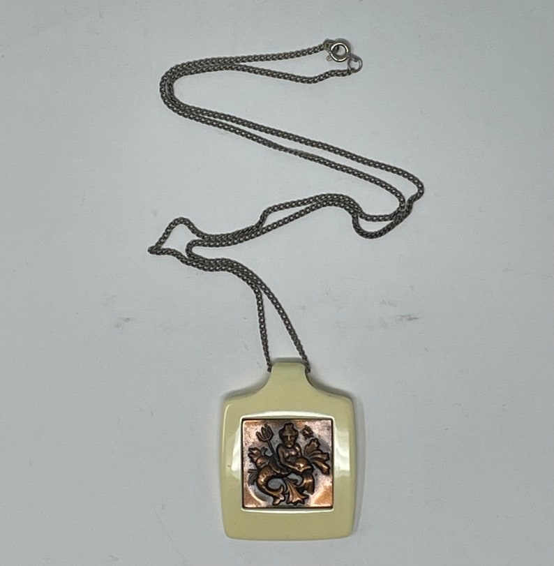 May include: A silver chain necklace with a square pendant. The pendant is white with a copper-colored image of a mermaid inside.