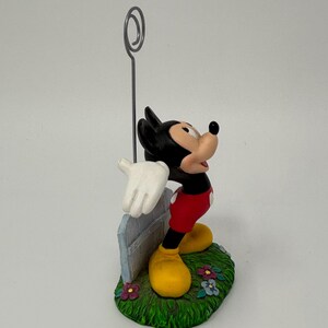 May include: A Mickey Mouse figurine with a silver wire photo holder. Mickey is wearing red shorts, yellow shoes, and white gloves. He stands on a green base with flowers and a light blue fence.