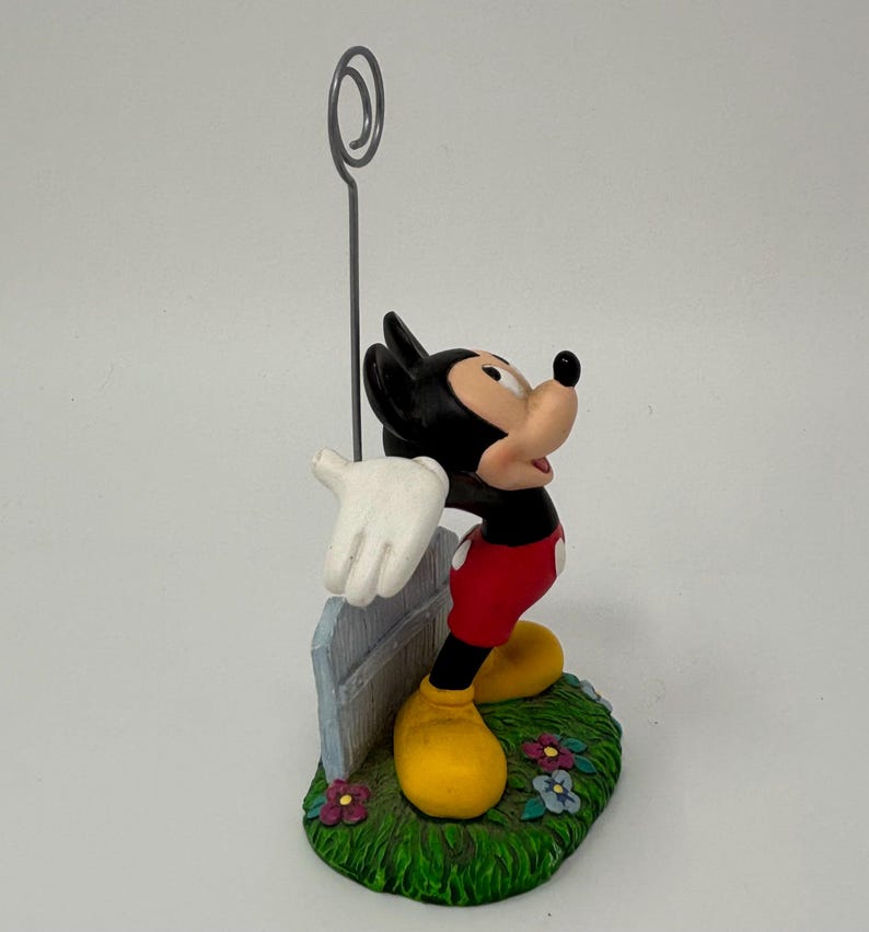 May include: A Mickey Mouse figurine with a silver wire photo holder. Mickey is wearing red shorts, yellow shoes, and white gloves. He stands on a green base with flowers and a light blue fence.