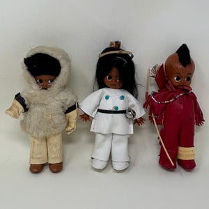 May include: Three vintage dolls dressed in traditional attire. One doll wears a white fur-lined parka, another a white outfit with turquoise accents, and the third a red outfit with a feathered headdress. Each doll has dark skin and painted facial features.