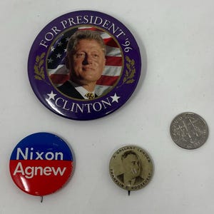 May include: A collection of political campaign buttons and a coin. The buttons feature portraits of former U.S. presidents and vice presidents, including Clinton, Nixon, Agnew, and Roosevelt. The buttons are round and colorful, with text and images.