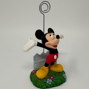 May include: A Mickey Mouse figurine with a silver wire photo holder. Mickey is in his classic outfit, standing on a grassy base with a small fence. The figurine has yellow shoes, red shorts, and white gloves.