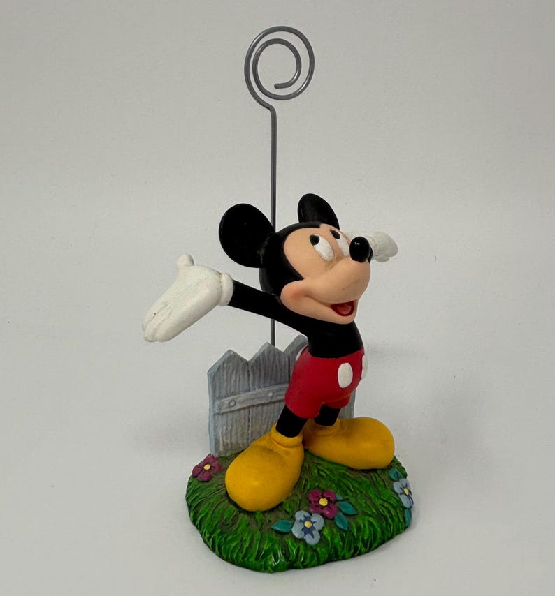 May include: A Mickey Mouse figurine with a silver wire photo holder. Mickey is in his classic outfit, standing on a grassy base with a small fence. The figurine has yellow shoes, red shorts, and white gloves.