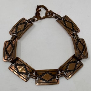 May include: A copper-toned bracelet with rectangular links, each featuring a geometric diamond design. The bracelet has a hook clasp and is likely vintage. The links are connected by small, rounded links.