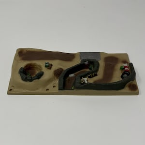 May include: A miniature toy battlefield diorama with a tan base, featuring a winding trench, a bunker, and small toy figures. The trench is dark green, and the base has brown and tan patches. Small toy vehicles and figures are visible.